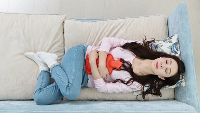 An Asian woman is lying on a sofa with a hot water bottle on her stomach